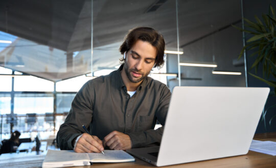 Busy young business man working in office elearning on laptop writing notes.