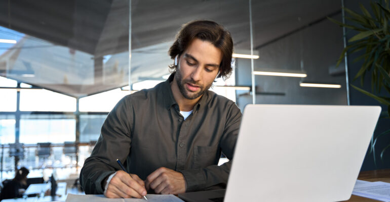 Busy young business man working in office elearning on laptop writing notes.