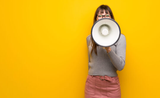 Woman with glasses over yellow wall shouting through a megaphone to announce something