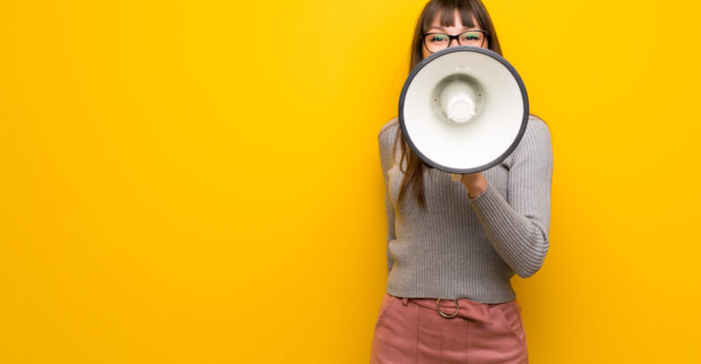 Woman with glasses over yellow wall shouting through a megaphone to announce something