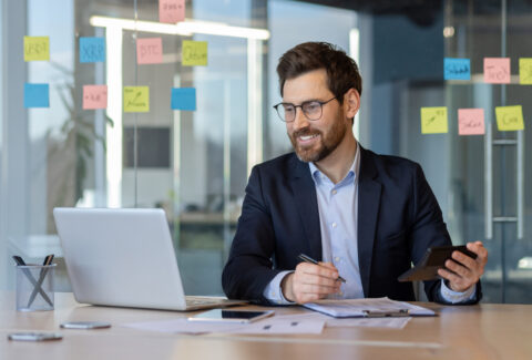 Businessman in modern office smiling while using calculator and laptop with sticky notes in background