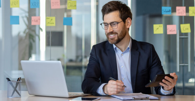 Businessman in modern office smiling while using calculator and laptop with sticky notes in background