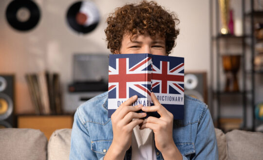 A teenager with curly hair sits focused in a room studying for an English exam. A boy in a blue jean shirt prepares for an English job interview